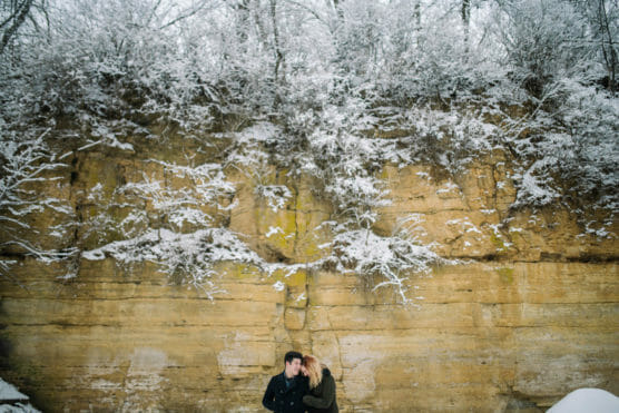 A couple stands close together in front of a tall cliff dusted with snow, with bare, snow-covered trees above the rock face. The scene feels wintry and intimate. by Justin Salem Meyer