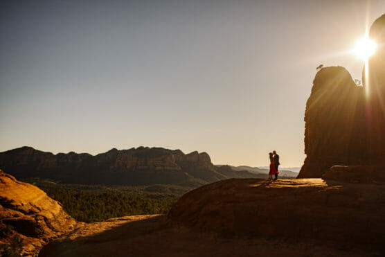 A couple stands embracing on a rocky ledge at sunset, surrounded by red rock formations and distant mountains under a clear sky, with the sun partially visible behind a large rock. by Justin Salem Meyer