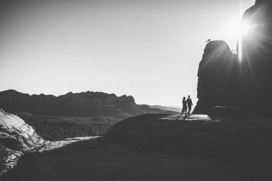 Two people stand together on a rocky landscape with mountains in the distance. The sun is shining behind a large rock, creating a dramatic, high-contrast black and white scene. by Justin Salem Meyer