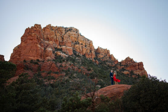 A couple stands together on a rocky ledge with the woman in a flowing red dress, surrounded by green shrubs and trees, against the backdrop of a towering red rock mountain under a clear sky. by Justin Salem Meyer