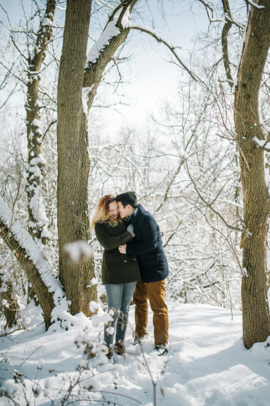 A couple stands in a snowy forest, embracing and smiling. They wear winter coats and boots, surrounded by tall trees dusted with snow, with a bright, clear sky above. by Justin Salem Meyer