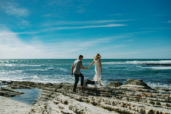 A couple walks hand in hand on rocky terrain by the ocean under a bright blue sky, with waves crashing in the background. by Justin Salem Meyer