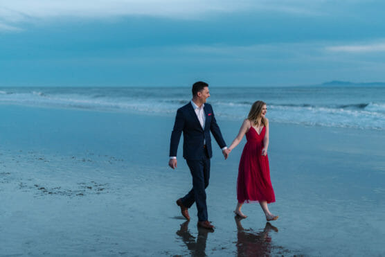 A man in a suit and a woman in a red dress walk hand in hand along a sandy beach at sunset, with gentle waves and a calm, blue sky in the background. by Justin Salem Meyer