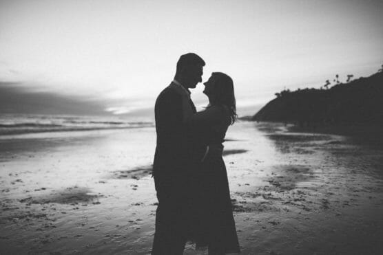 Silhouette of a couple standing close together on a beach at sunset, facing each other with the ocean and a distant hill in the background. The image is in black and white. by Justin Salem Meyer