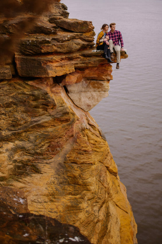 A couple sits together on the edge of a high, rocky cliff overlooking a body of water, with layers of orange and yellow stone visible beneath them. by Justin Salem Meyer