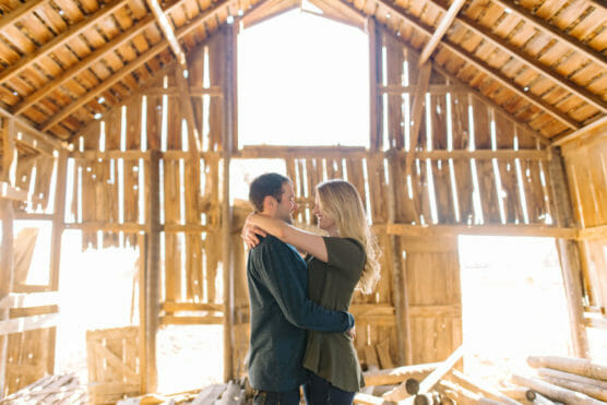 A couple stands inside a sunlit, rustic wooden barn, embracing each other and gazing into each others eyes, surrounded by scattered wooden planks and beams. by Justin Salem Meyer