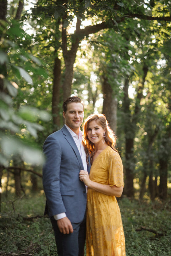 A couple stands closely together in a sunlit forest. The man wears a blue suit and the woman wears a yellow dress. They both smile at the camera, surrounded by lush green trees and soft, natural light. by Justin Salem Meyer