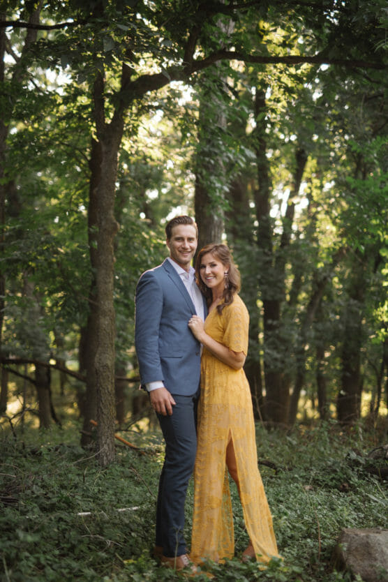 A smiling couple stands together in a sunlit forest. The man wears a blue suit, and the woman wears a long yellow dress. They are close, with trees and greenery surrounding them. by Justin Salem Meyer