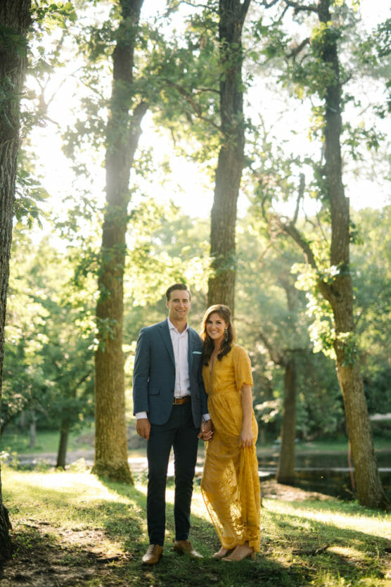A couple stands together in a sunlit forest clearing, holding hands and smiling. The man wears a blue suit and brown shoes; the woman wears a yellow lace dress. Sunlight filters through tall trees behind them. by Justin Salem Meyer