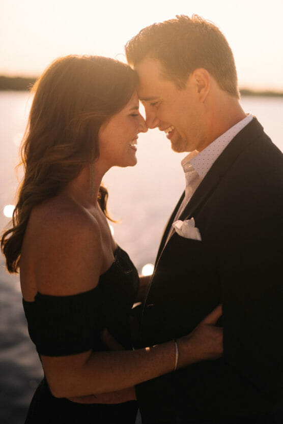A couple stands close together at sunset by the water, smiling with their foreheads touching. The woman wears an off-the-shoulder dress, and the man is dressed in a suit. The warm sunlight creates a romantic and intimate atmosphere. by Justin Salem Meyer