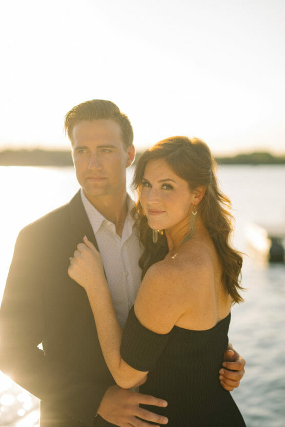 A couple stands close together by the water at sunset. The woman, wearing a black off-shoulder dress and earrings, has her arm around the mans shoulder. The man is in a suit, looking into the distance with a soft, warm glow behind them. by Justin Salem Meyer