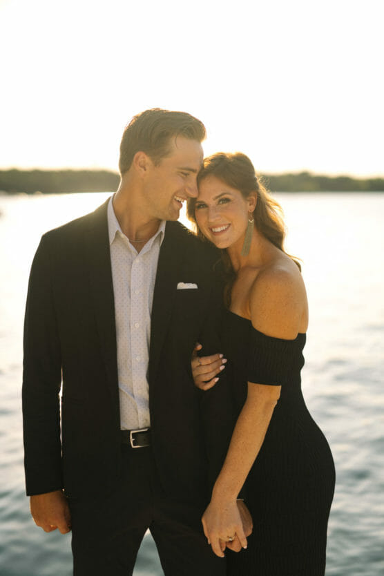 A couple dressed formally, standing close together and smiling by a sunlit body of water. The man wears a dark suit and the woman wears an off-the-shoulder black dress with earrings, both appearing happy and relaxed. by Justin Salem Meyer