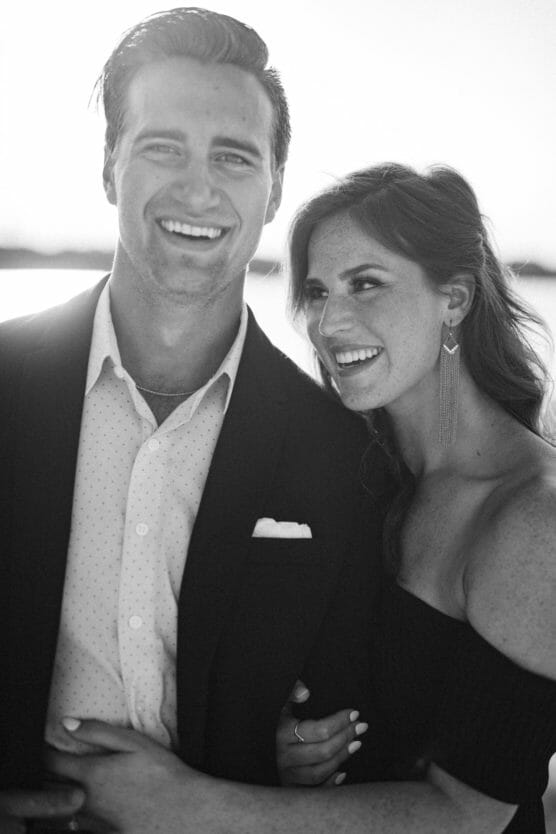 A black-and-white photo of a smiling couple. The man wears a suit with a pocket square and an open-collar shirt, while the woman, in a strapless dress, stands beside him with her arm around his, both looking happy. by Justin Salem Meyer