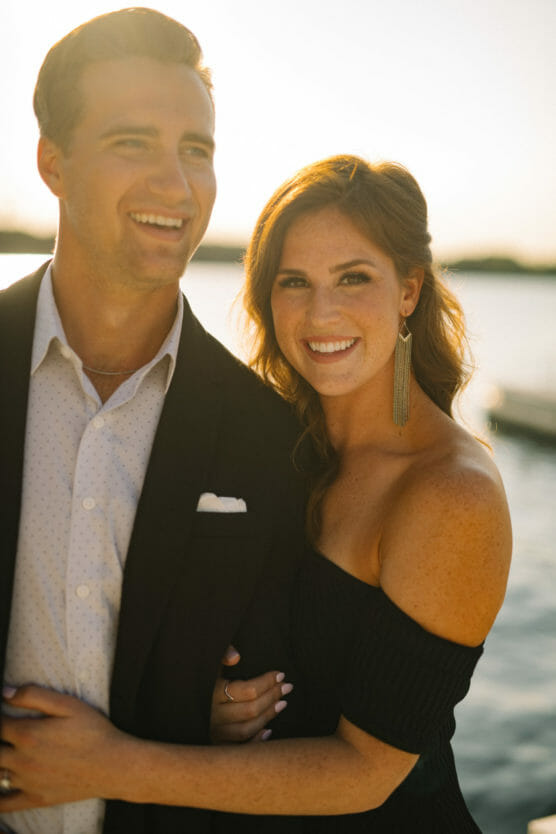 A smiling couple poses together outdoors near water at sunset. The man wears a black suit and white shirt, while the woman wears a black off-shoulder dress and long earrings, with her arm around him. by Justin Salem Meyer
