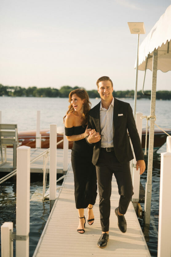 A smiling couple dressed in formal attire walks hand in hand on a dock by the water, with boats and trees in the background on a sunny day. by Justin Salem Meyer