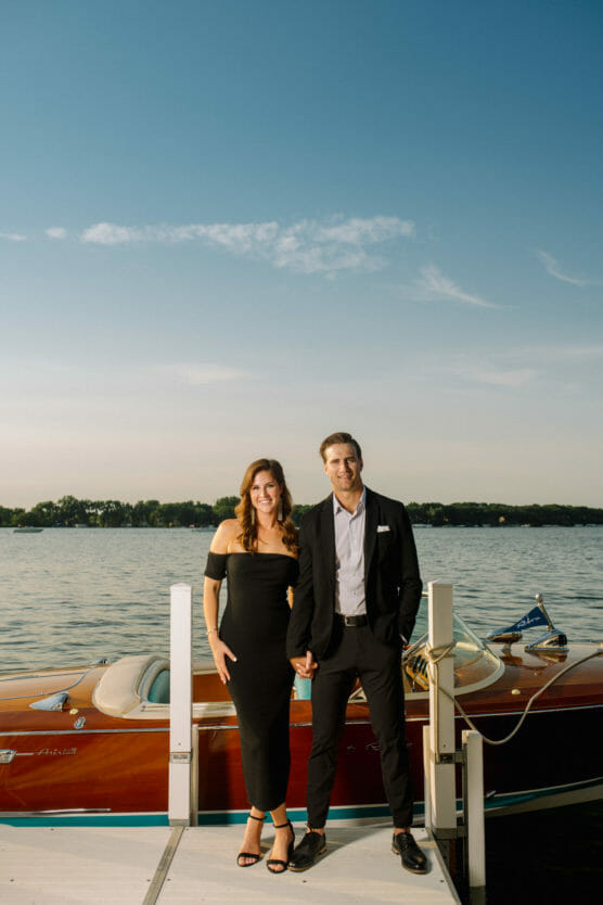 A couple dressed formally, with the woman in a black dress and the man in a black suit, stand smiling on a dock by a wooden boat with a lake and blue sky in the background. by Justin Salem Meyer