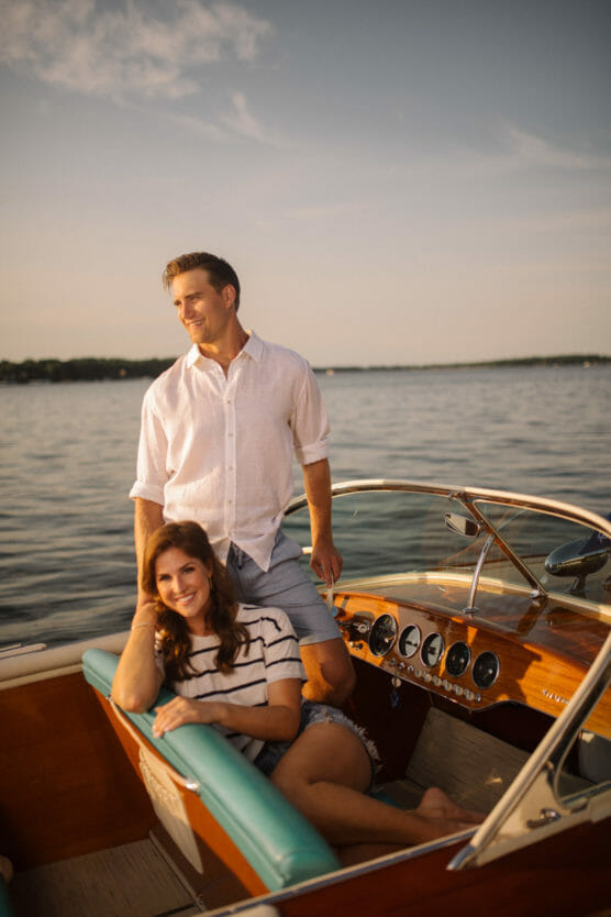 A man in a white shirt stands at the helm of a wooden boat, while a woman in a striped shirt and shorts sits smiling on a green seat. They are on calm water under a clear sky at sunset. by Justin Salem Meyer