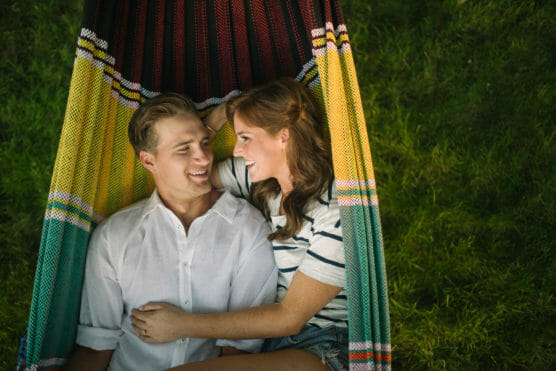 A couple lies together in a colorful hammock, smiling at each other on a sunny day. The woman has her arm around the man, and they are surrounded by green grass. by Justin Salem Meyer