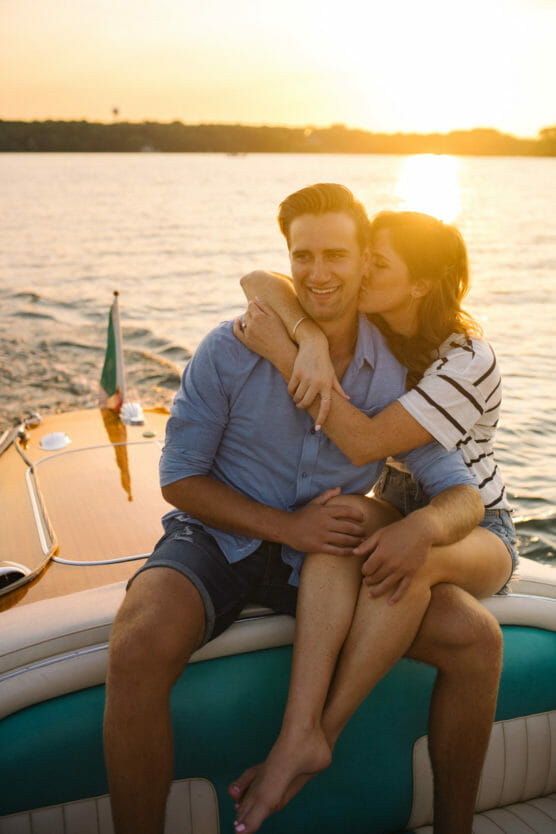 A woman hugs and kisses a smiling man on a boat at sunset. They both appear happy and relaxed, sitting close together with water and the sun in the background. by Justin Salem Meyer