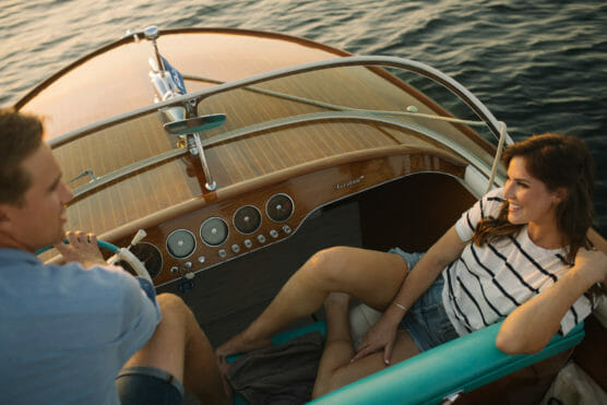 A man steers a wooden boat while a woman in a striped shirt and denim shorts relaxes on a turquoise seat, smiling, as they travel on calm water at sunset. by Justin Salem Meyer