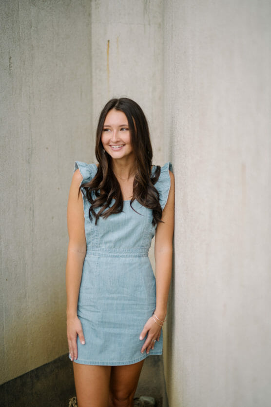 A young woman with long brown hair, wearing a light blue denim dress, stands and smiles while leaning against a light-colored textured wall. by Justin Salem Meyer