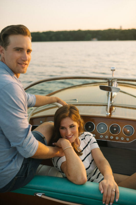 A man and a woman relax on a boat at sunset. The man sits on the edge near the steering wheel, while the woman leans back smiling on the seat in front of him. Calm water and green trees are visible in the background. by Justin Salem Meyer