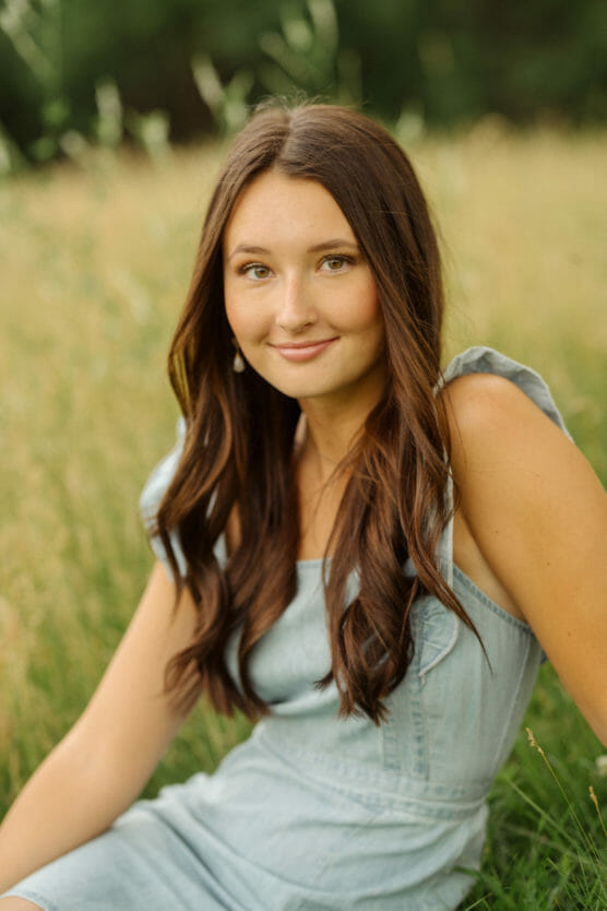 A young woman with long brown hair sits in a grassy field, wearing a light blue sleeveless dress. She is smiling softly and looking at the camera, with greenery and tall grass in the blurred background. by Justin Salem Meyer