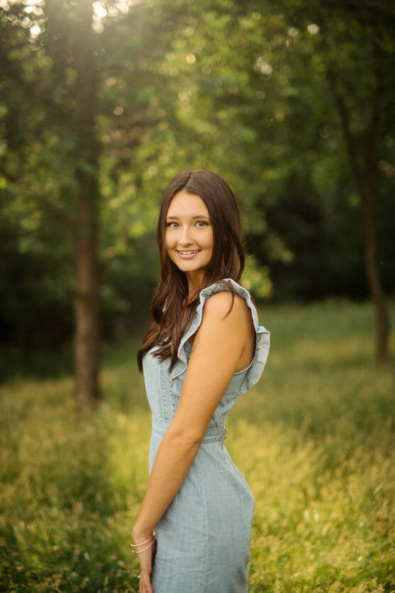 A young woman with long brown hair, wearing a sleeveless light blue dress, stands smiling in a sunlit, grassy area with trees in the background. by Justin Salem Meyer