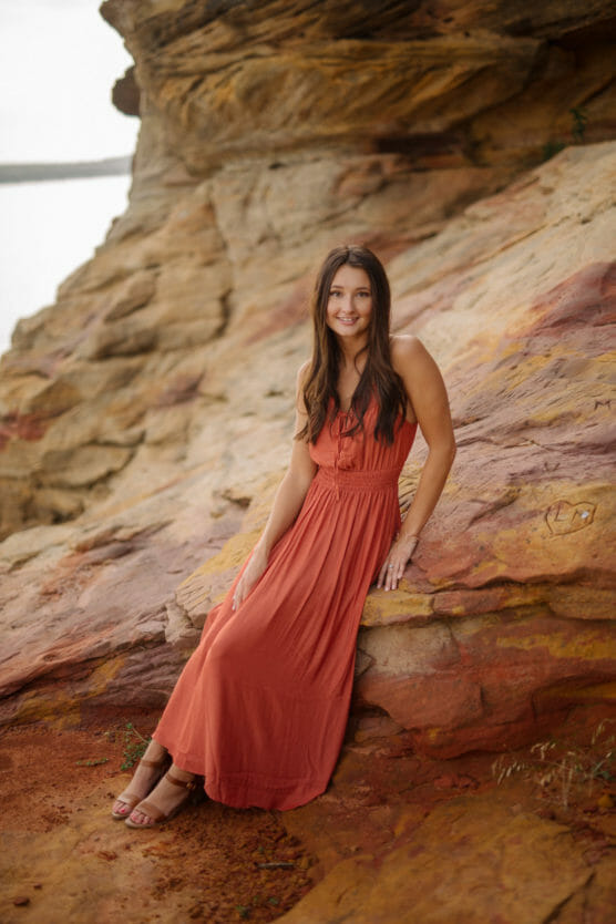 A woman in a flowing rust-colored dress sits on a large, multi-colored rock formation outdoors, smiling at the camera. The background features textured rock layers and soft natural light. by Justin Salem Meyer