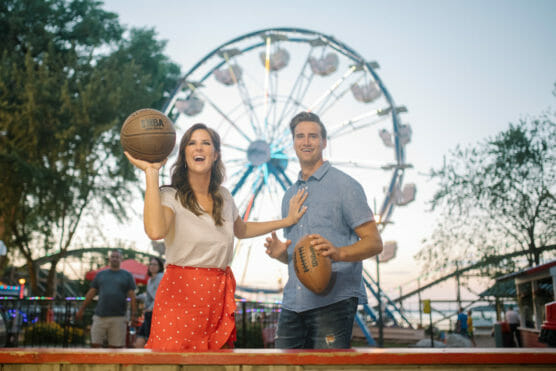 A smiling woman holding a basketball prepares to throw it at a carnival game, while a man beside her holds another basketball. A Ferris wheel and trees are visible in the background. by Justin Salem Meyer
