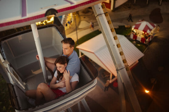 A young couple sits closely together in a Ferris wheel gondola at dusk, with the amusement park and carnival booths visible below them. The scene is warm and intimate, viewed from above. by Justin Salem Meyer