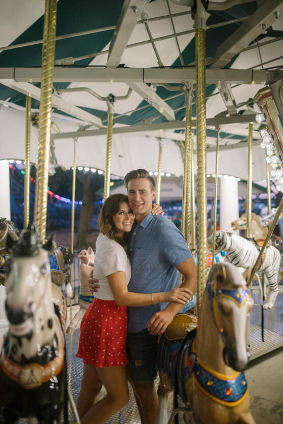 A couple smiles and hugs while standing on a carousel, surrounded by colorful carousel horses and festive lights. The woman wears a white top and red skirt, and the man wears a blue shirt and dark shorts. by Justin Salem Meyer