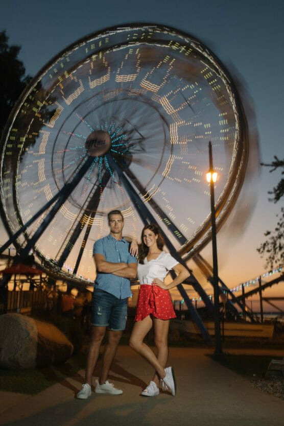 A man and woman stand smiling in front of a brightly lit, spinning Ferris wheel at dusk. The man is in a blue shirt and shorts with arms crossed; the woman wears a white top and red skirt with hands on hips. by Justin Salem Meyer
