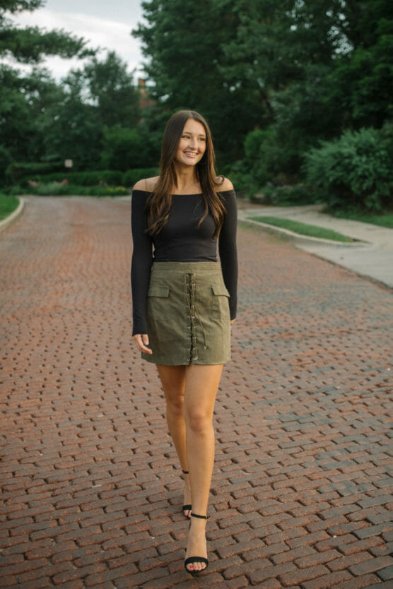 A woman with long brown hair walks on a brick street, wearing a black off-the-shoulder top, an olive green skirt with buttons, and black heeled sandals. Trees and greenery line the background. by Justin Salem Meyer