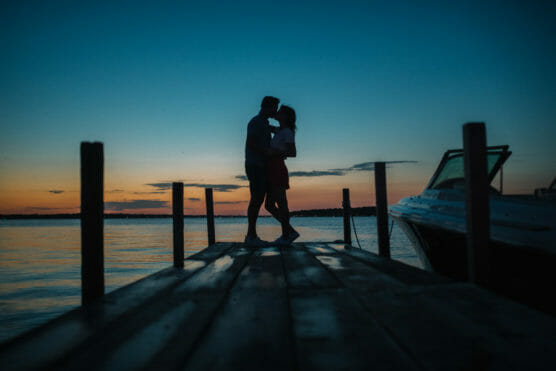 Silhouette of a couple embracing on a wooden dock at sunset, with a boat to the right and calm water in the background. The sky is a gradient of blue and orange. by Justin Salem Meyer