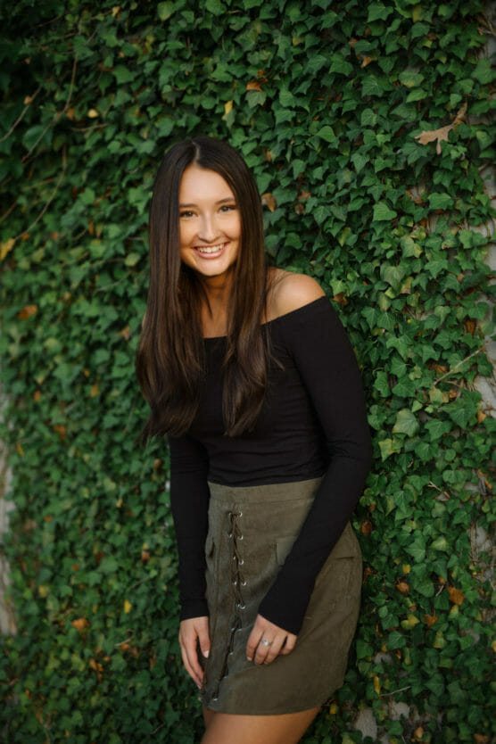 A young woman with long brown hair, wearing an off-the-shoulder black top and olive green skirt, stands smiling in front of a wall covered with green ivy. by Justin Salem Meyer