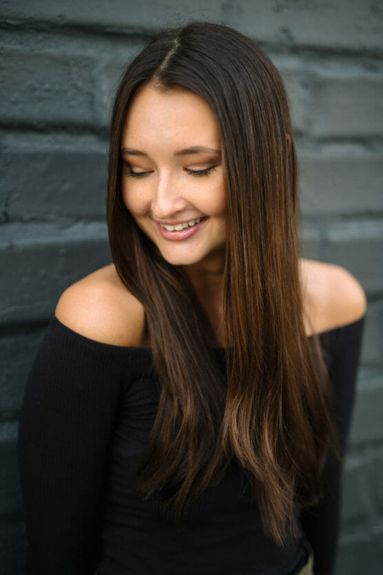 A young woman with long brown hair, wearing a black off-the-shoulder top, stands against a dark gray brick wall and looks down with a gentle smile. by Justin Salem Meyer