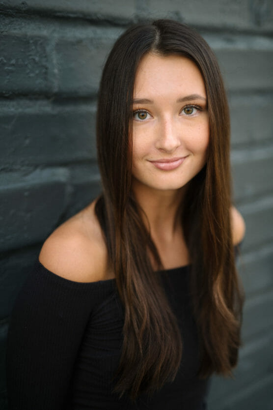 A young woman with long brown hair and hazel eyes smiles softly, wearing a black off-the-shoulder top, standing in front of a dark gray brick wall. by Justin Salem Meyer