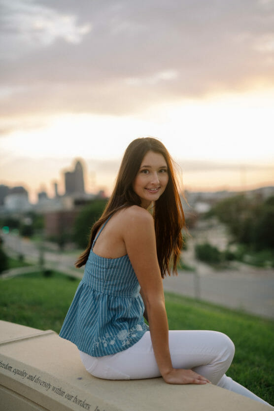 A young woman with long brown hair, wearing a blue sleeveless top and white pants, sits on a ledge outdoors at sunset with a city skyline in the background, smiling at the camera. by Justin Salem Meyer