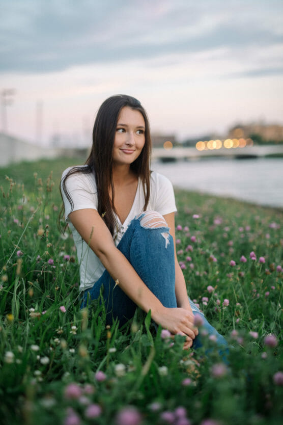 A young woman with long brown hair sits in a field of green grass and pink clover flowers, wearing a white t-shirt and ripped blue jeans, smiling softly at sunset near a river. by Justin Salem Meyer