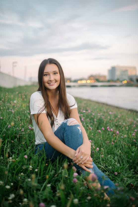 A young woman with long brown hair sits on green grass with pink clover flowers by a river, smiling at the camera. She wears a white shirt and ripped blue jeans, with city buildings blurred in the background. by Justin Salem Meyer