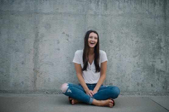 A young woman with long dark hair sits cross-legged on the ground in front of a concrete wall, smiling and laughing, wearing a white shirt and ripped blue jeans. by Justin Salem Meyer