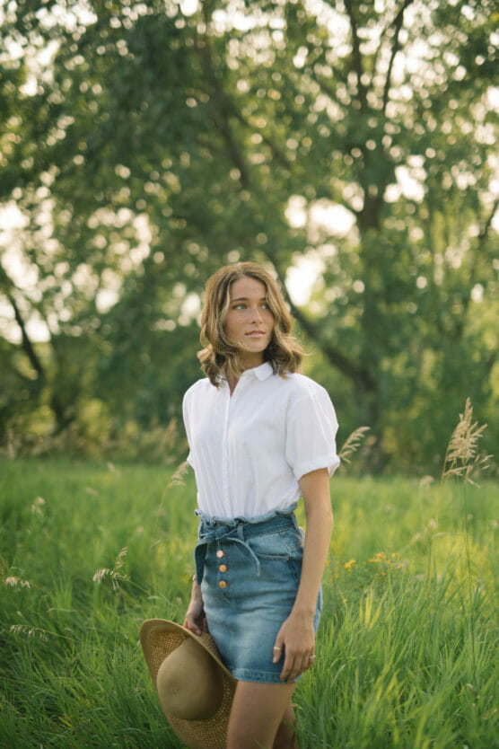 A woman stands in a grassy field holding a straw hat by her side. She wears a white short-sleeve shirt and a denim skirt, with trees and soft sunlight in the background. by Justin Salem Meyer