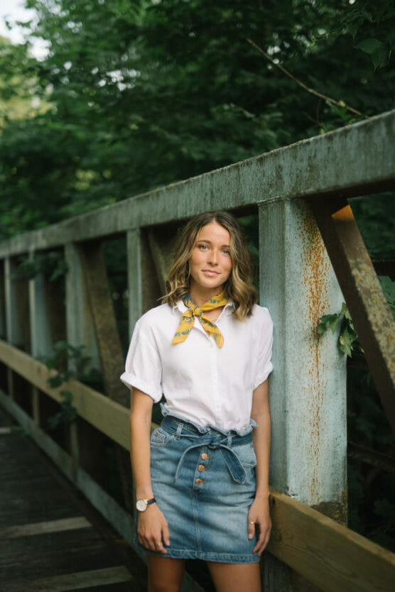 A young woman stands on a metal bridge surrounded by greenery. She is wearing a white shirt, denim skirt, yellow neck scarf, and a watch, with her hands by her side, looking at the camera and smiling slightly. by Justin Salem Meyer