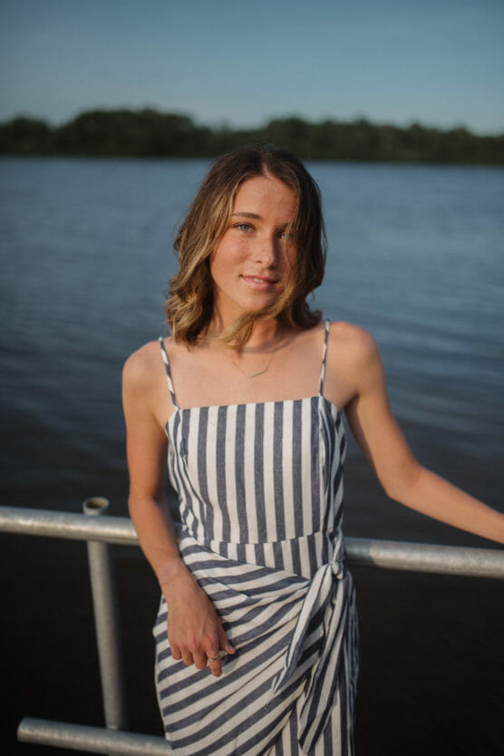 A young woman with wavy brown hair stands by a railing in front of a calm lake, wearing a sleeveless, black and white striped dress, with trees and blue sky in the background. by Justin Salem Meyer