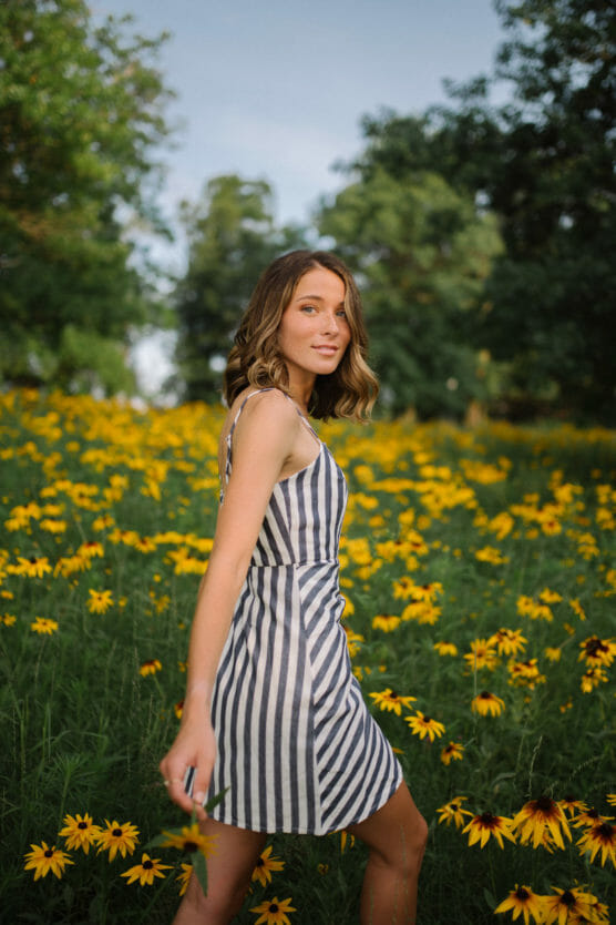 A young woman in a black-and-white striped dress stands in a field of yellow flowers, with green trees in the background. She looks toward the camera, one arm slightly raised, under a blue sky. by Justin Salem Meyer
