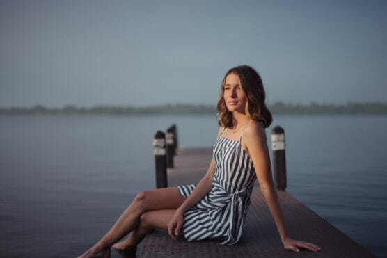 A young woman in a sleeveless, striped dress sits on a wooden dock by calm water, with a misty background and soft morning or evening light. by Justin Salem Meyer