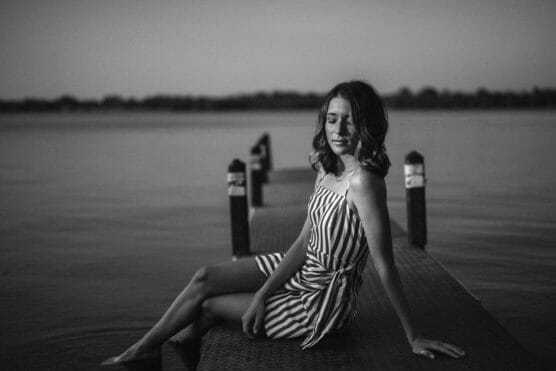 A young woman in a striped dress sits on a dock by calm water, eyes closed and head tilted slightly, with trees visible in the distant background. The photo is in black and white. by Justin Salem Meyer