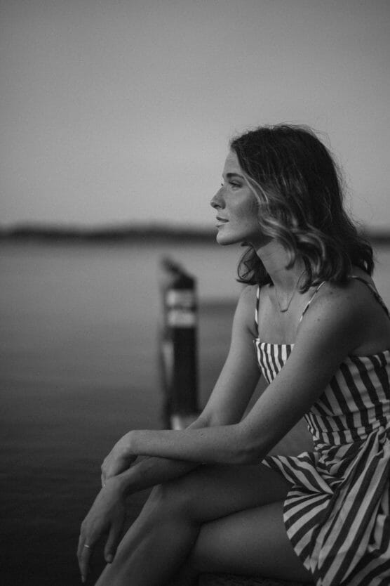 A woman in a striped dress sits by the water, gazing thoughtfully into the distance. The black-and-white photo creates a calm and reflective mood. by Justin Salem Meyer