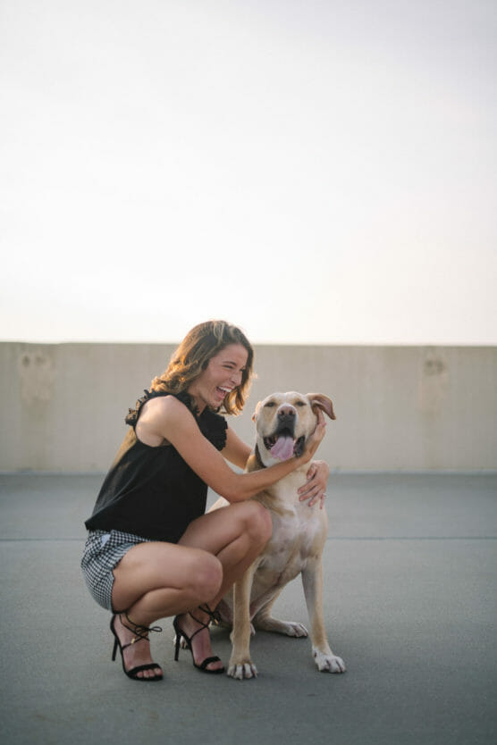 A woman in a black sleeveless top, checked shorts, and heels kneels on concrete, smiling and hugging a happy, tan dog with a white chest. The background is an empty outdoor area with a light sky. by Justin Salem Meyer